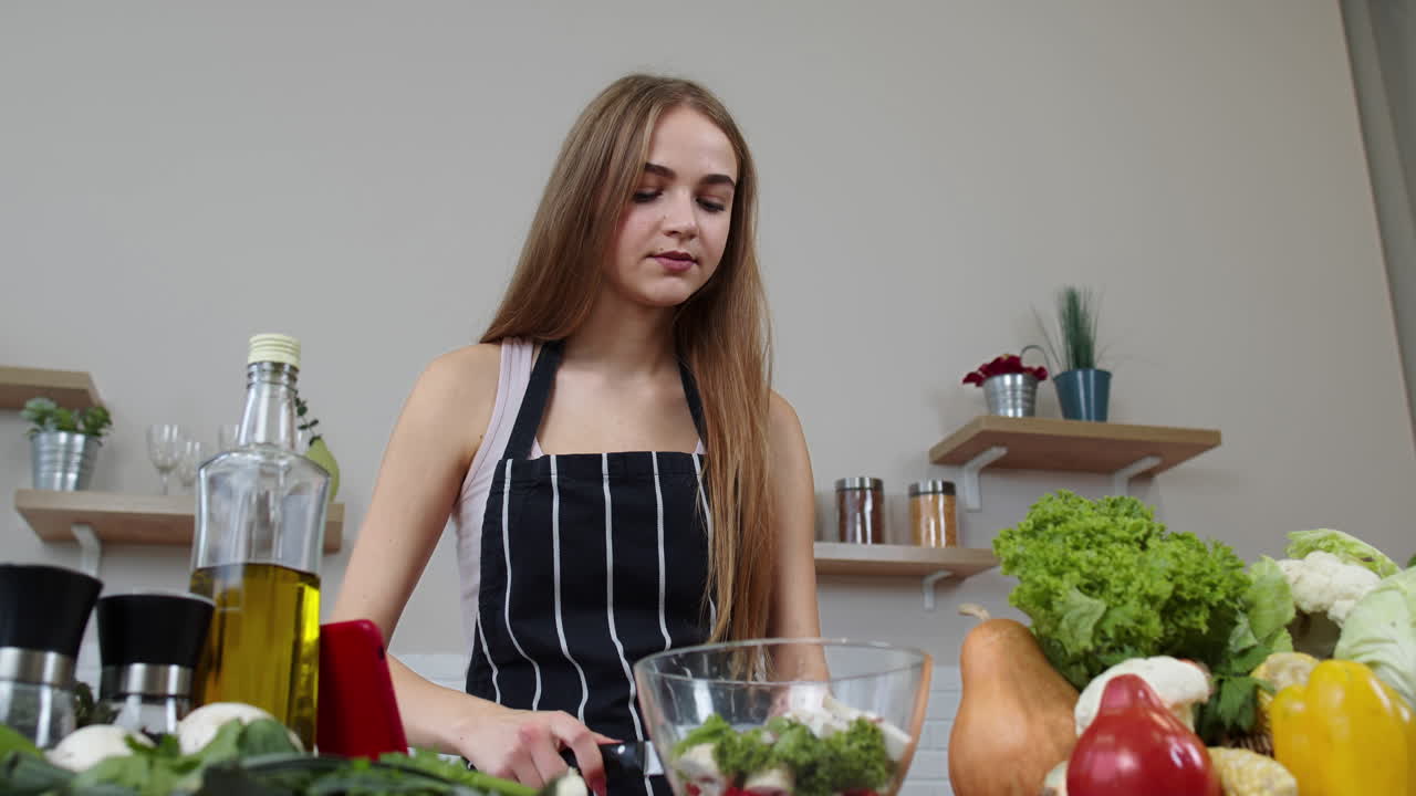 Niña vegana cocinando ensalada con verduras crudas mientras busca receta en línea en su teléfono móvil
