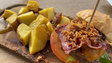 mini hamburguesa con tocino, cebollas crujientes, tomate, lechuga y cubos de patata al lado en un restaurante en un plato de madera, 4k paneo a la izquierda