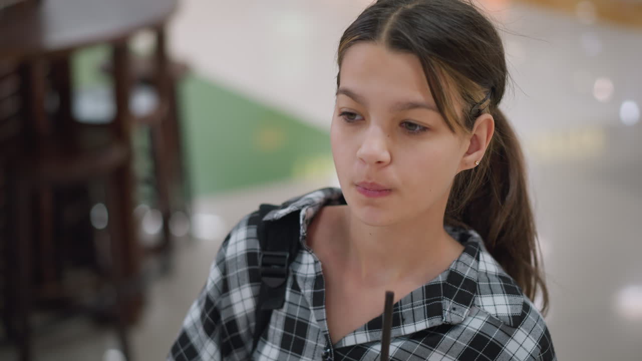 세련된 카레드 셔츠를 입은 어린 소녀는 을 통해 주스를 마시면서 고, 그녀는 배을 들고, 평온한 표정으로 내려다보고, 흐릿한 배경은 의자와 실내 조명을 특징으로합니다.