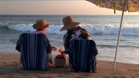 an elderly couple share drinks under an umbrella on the beach