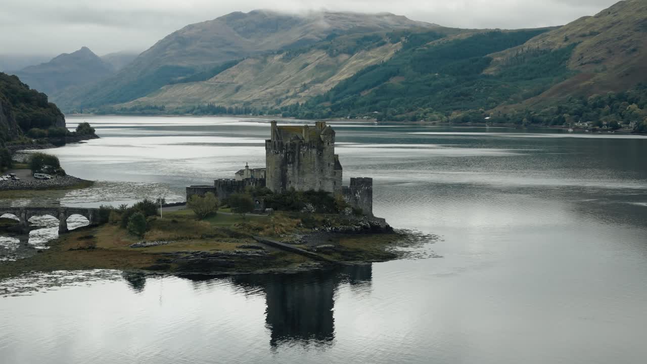 Retreating aerial circling shot of Eilean Donan Castle reflecting on calm lake