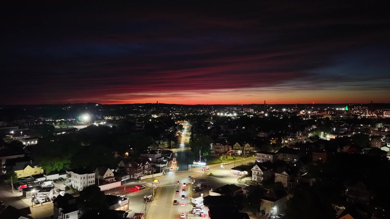 Strade urbane illuminate dai lampioni sotto un profondo bagliore crepuscolare cremisi, tetti scuri e incroci illuminati che si estendono verso le luci della città in lontananza di notte. Massachusetts, USA. Sorvolo aereo.