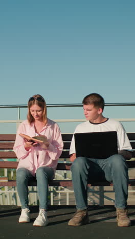 estudiantes masculinos y femeninos sentados al aire libre en un banco estudiando juntos, el niño escribe en la computadora portátil y escucha mientras la niña lee de un libro, asintiendo con la cabeza en acuerdo, en un telón de fondo urbano