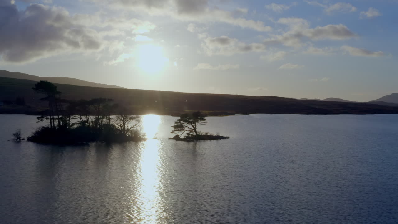 Aerial tracking right across Lough Bofin in Ireland, small tree covered islands under golden evening light backlit, Connemara, Galway, Ireland