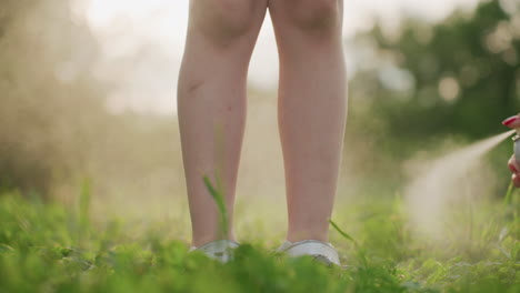 close up of leg view of girly figure standing on grass while hand sprays mist on her leg, summer park setting with warm light, outdoor child protection and cooling during playful family leisure