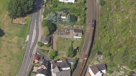 drone shot offering a panoramic view of a quaint small town, interwoven with a bustling railway line and road, accentuated by charming white buildings with red roofs, surrounded by lush greenery