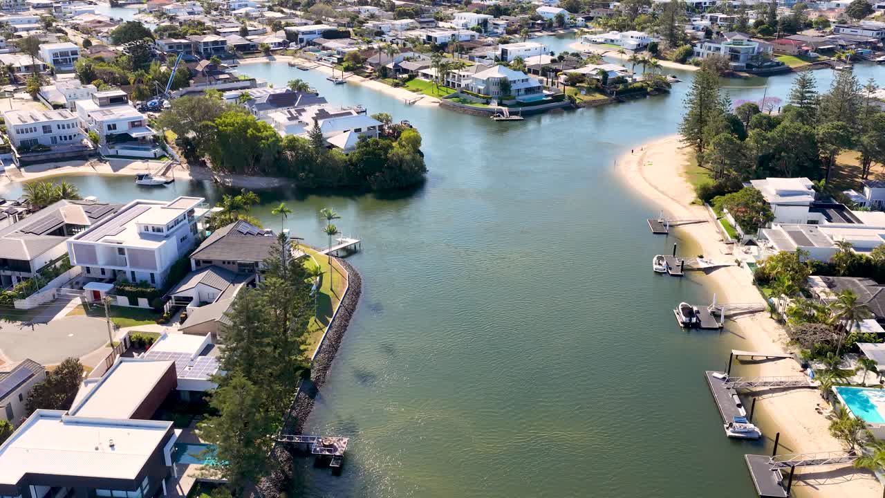 Drone glides above riverside residential neighborhood, modern houses, docks, and waterways under bright sunlight