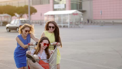 friends entertaining in mall parking lot in bright day. energetic women circling attractive cheerful girl in shopping cart enjoying free time, slomo