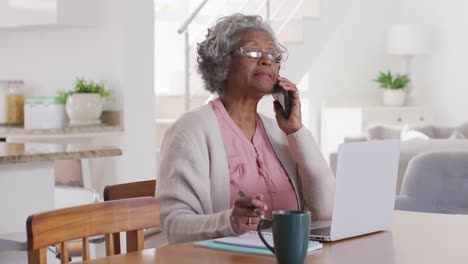 Portrait-of-senior-african-american-woman-sitting-at-table,-using-laptop-and-talking-on-smartphone