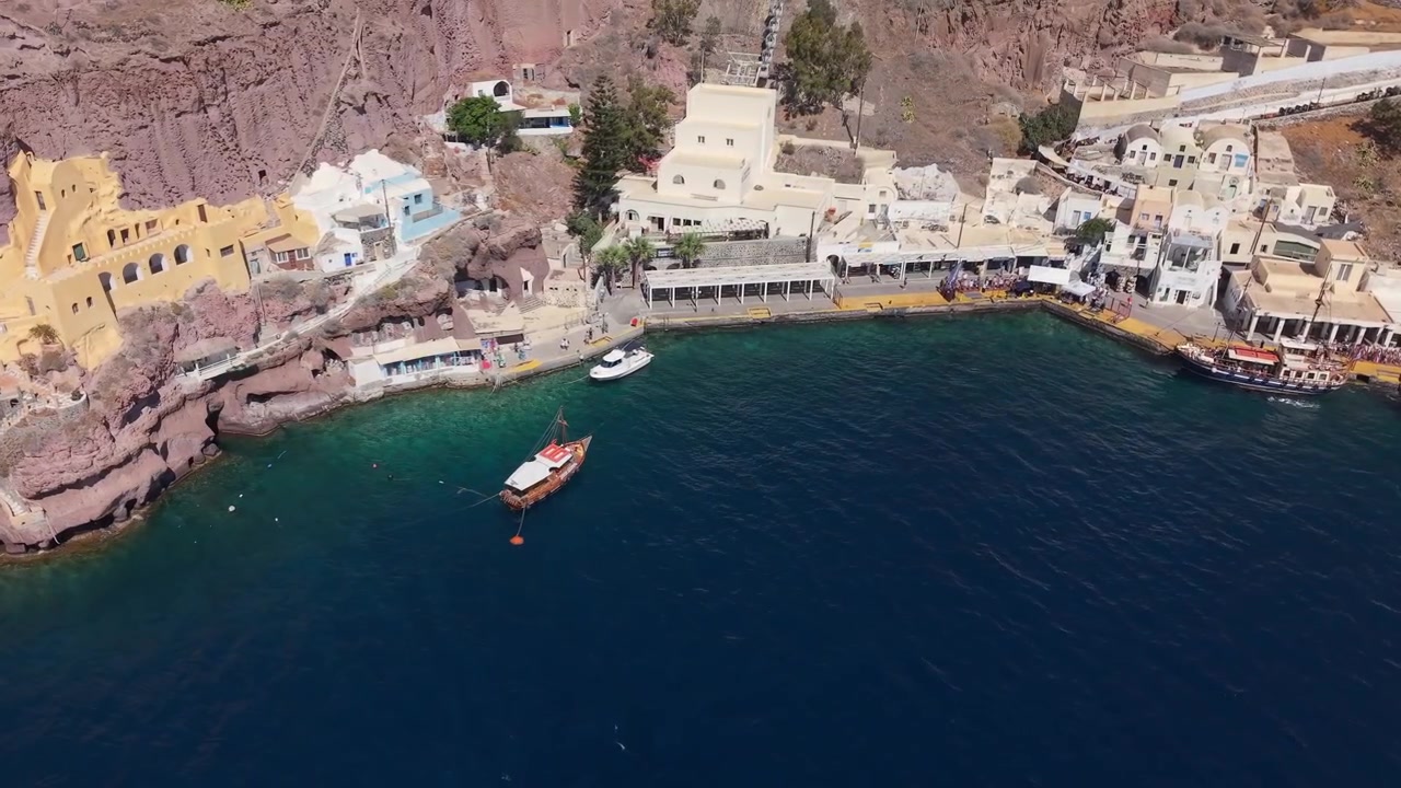 Drönarvy över Santorinis gamla hamn som avslöjar ett unikt vulkaniskt kalderalandskap, båtar i hamnen och de dramatiska klipporna nedanför Fira.