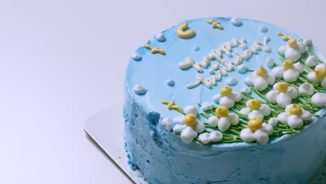 Close-up-shot-of-the-top-of-a-birthday-cake-with-blue-frosting-and-decorated-with-white-and-yellow-frosted-flowers,-with-happy-birthday-greetings-on-top-of-it