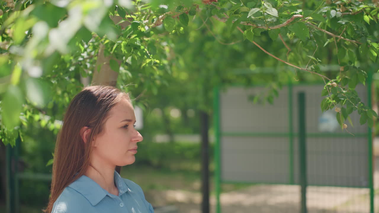 코카서스 인종 여성이 울타리 근처 통로에서 옆모습을 보이며 공원을 가로질러 생각에 잠긴 듯한 시선을 보내고 있어요. 캐주얼한 데님 셔츠를 입고, 나뭇잎이 우거진 배경에 조용한 여름 분위기이며, 사색에 잠긴 자세로 도시와 자연이 어우러져 있습니다.