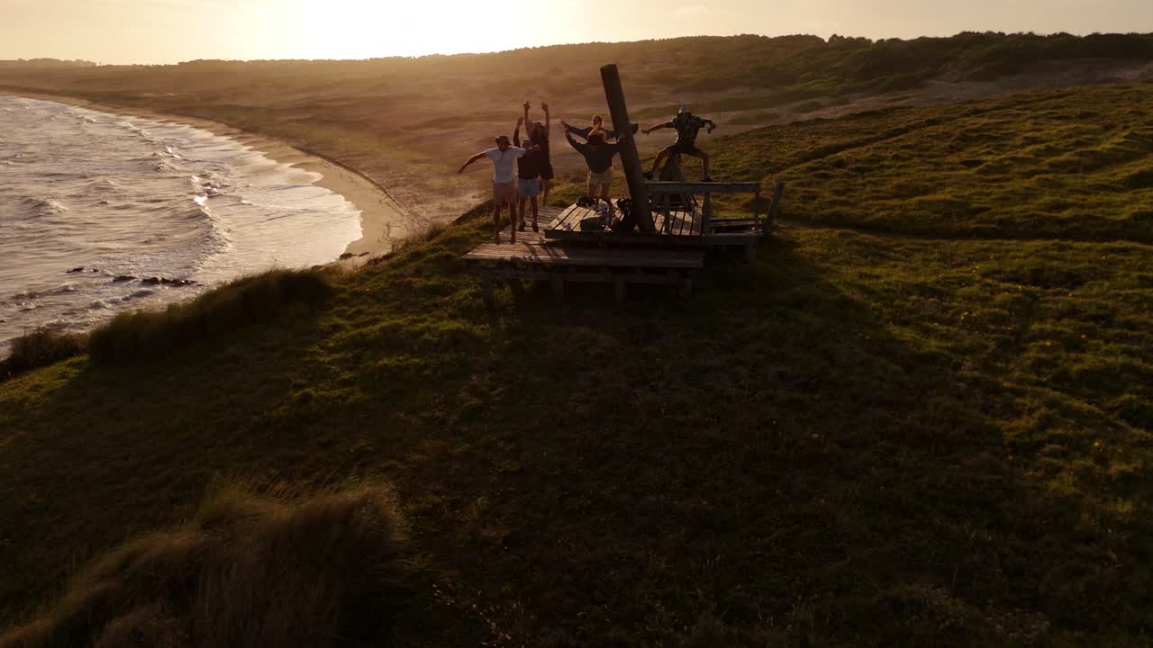 Seis amigos felizes pulando e celebrando juntos em um mirante no Cerro Chato, olhando para a câmera durante um pôr do sol dourado no Uruguai. Em câmera lenta.