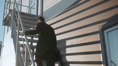 hombre de negocios en abrigo negro subiendo la escalera sosteniendo una barandilla de hierro y un maletín, con la luz del sol creando sombras dramáticas en la pared del edificio moderno a rayas y el reflejo de la cúpula en la ventana de vidrio