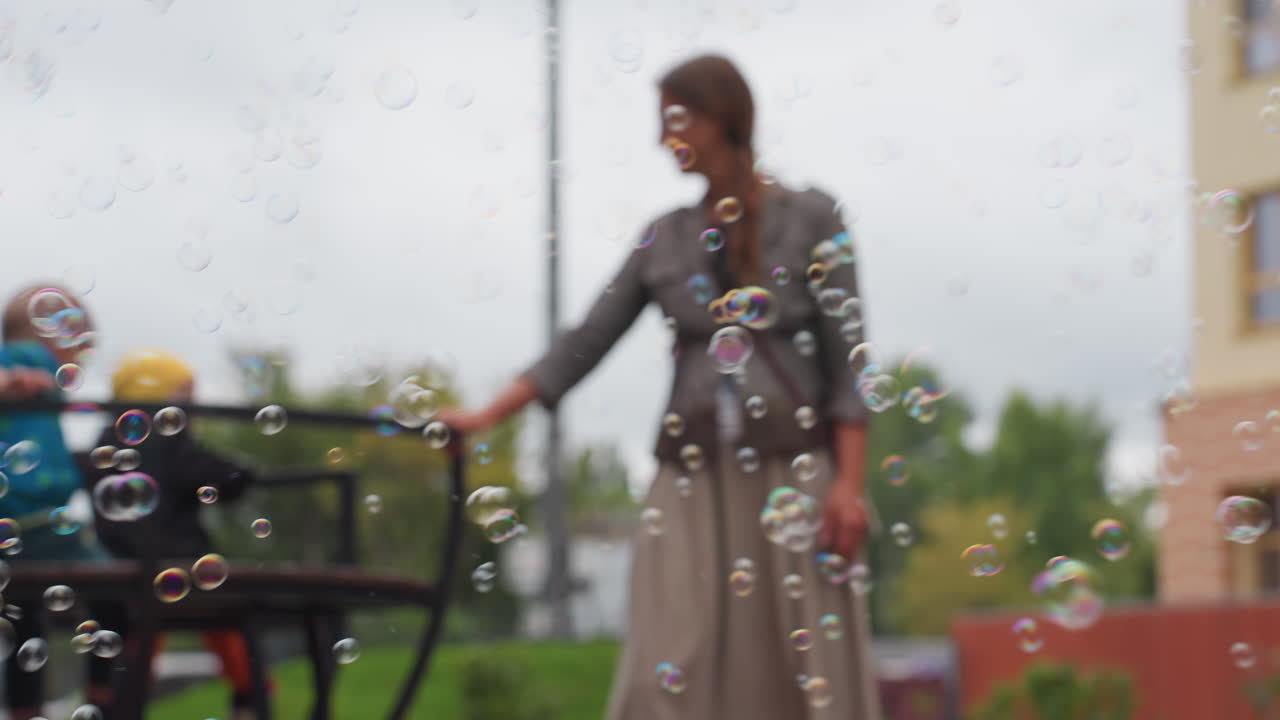 Caregiver hold merry go round children seated on playground surrounded by floating bubbles under cloudy sky, joyful outdoor moment with soft focus foreground
