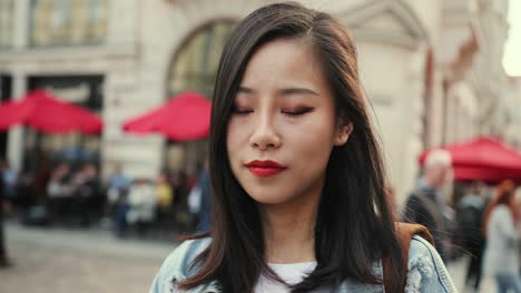 Mujer-Joven-Y-Guapa-Con-Labios-Rojos-Mirando-Directamente-A-La-Cámara-Y-Sonriendo-A-La-Plaza-De-La-Ciudad-Con-Sombrillas-De-Cafés