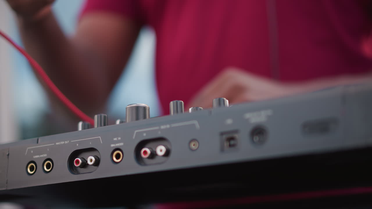 Siyah bir DJ mikserde kablo bağlantısı yapıyor, yakın çekim eller kırmızı RCA'yı girişe takıyor, kazanç düğmesini ayarlıyor ve sinyali test ediyor, odaklanmış müzisyen canlı setine hazırlanırken kentsel çatı katında ses kontrolü