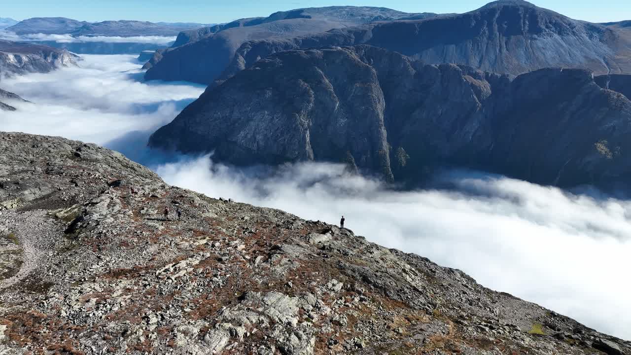 Drone starts close to a person, then reverses and tilts up, revealing Naeroyfjord, and the valley. Three others walk away, with the landscape unfolding