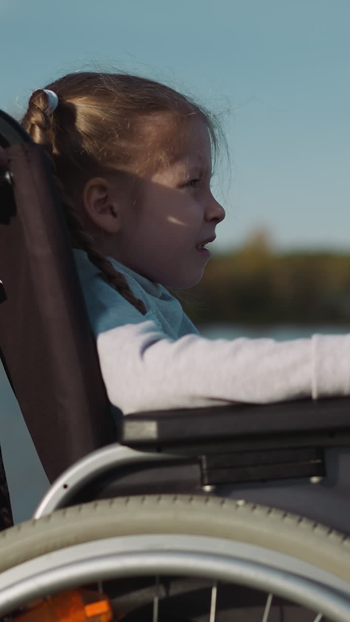 Preschooler girl with braids sits in wheelchair on coast of lake. Caring young mother stands near daughter resting with kid on blurred background closeup side view