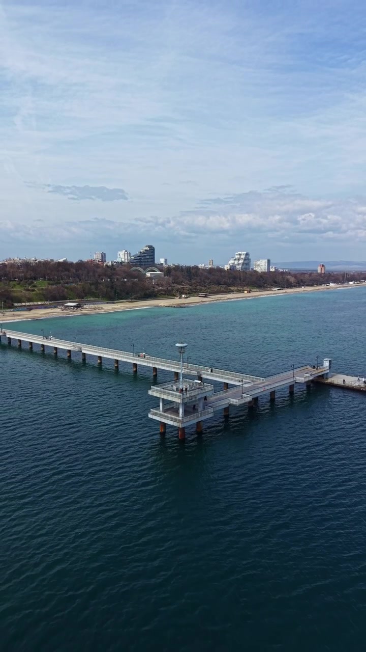 Vista di un grande molo con persone sulla sua struttura, sull'acqua, di giorno, vicino a una città e una spiaggia con nuvole nel cielo.