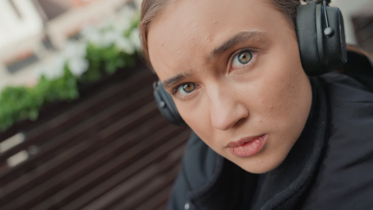 young woman in headphones looks hopeful on outdoor bench, soft city terrace and wooden slats, attentive gaze and relaxed posture, subtle freckles and natural light, roles include traveler, creator