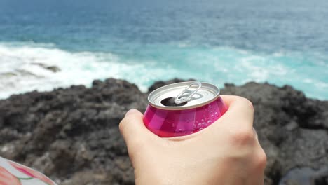 Close-up-of-hand-holding-refreshing-red-can-beverage-while-admiring-sea-coast