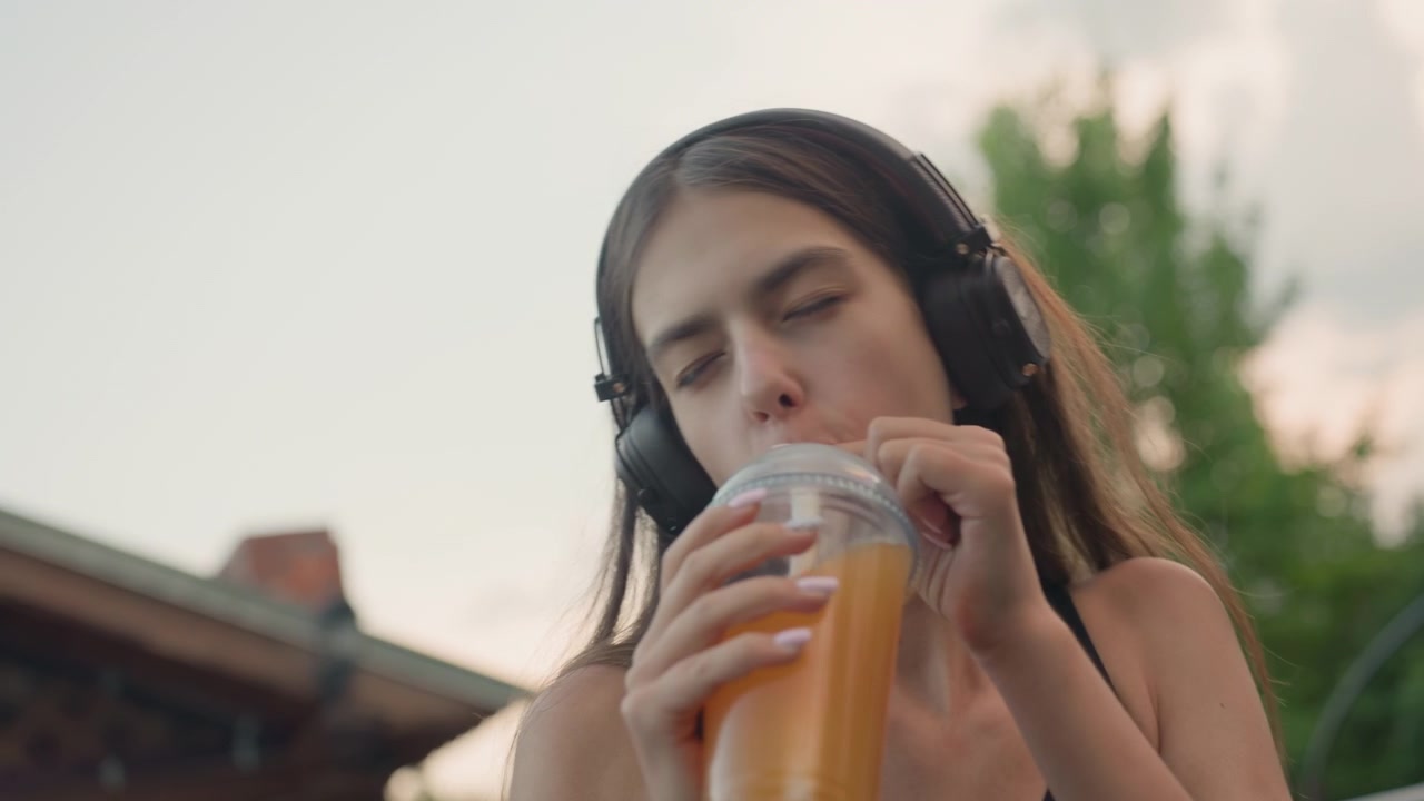 Giovane donna si rilassa all'aperto bevendo succo d'arancia mentre indossa le cuffie. La donna è seduta vicino a una casa di mattoni con vegetazione sullo sfondo, sotto una luce naturale soffusa.