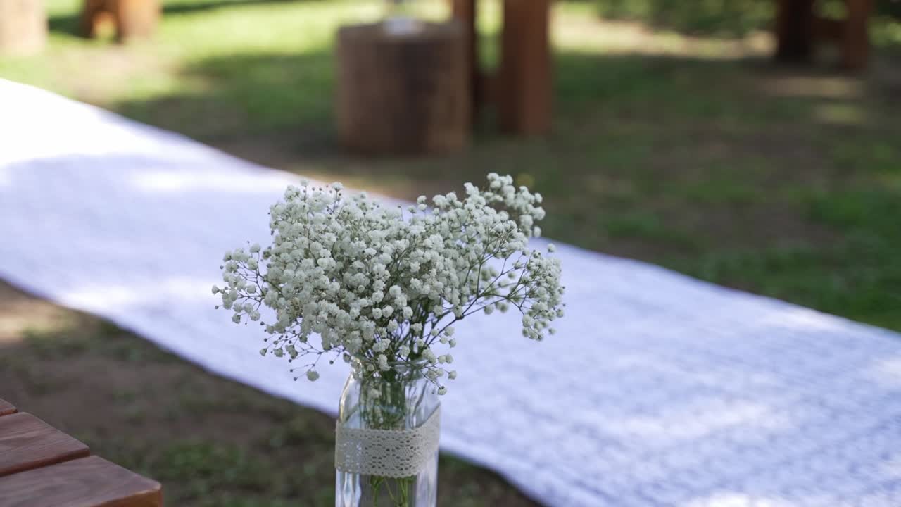 Delicato bouquet di gipsofila in un semplice vaso di vetro che decora un corridoio per cerimonia all'aperto con tappeto bianco, creando un'atmosfera morbida, romantica e pacifica.