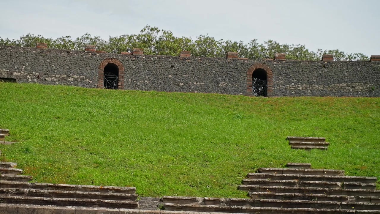 Gradini in pietra e arena erbosa all'interno dell'Anfiteatro di Pompei, con cancelli ad arco, antiche mura e sedili romani conservati sotto una luce diurna soffusa.
