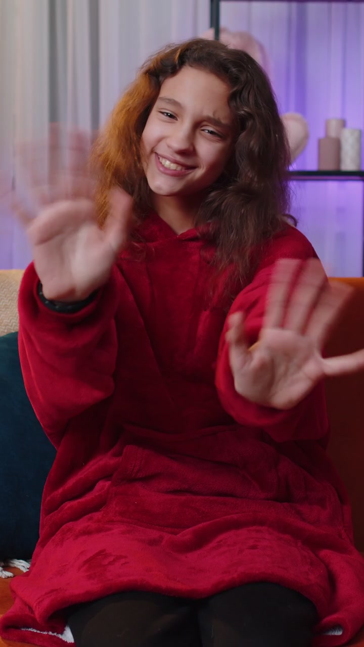 Ragazza preadolescente sorridente che guarda amichevolmente la fotocamera, salutando con la mano a casa sul divano.