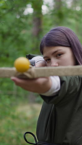 teenage girl with long purple hair in medieval dress aims in camera with crossbow in green forest. role-player at quest. acting and reconstruction