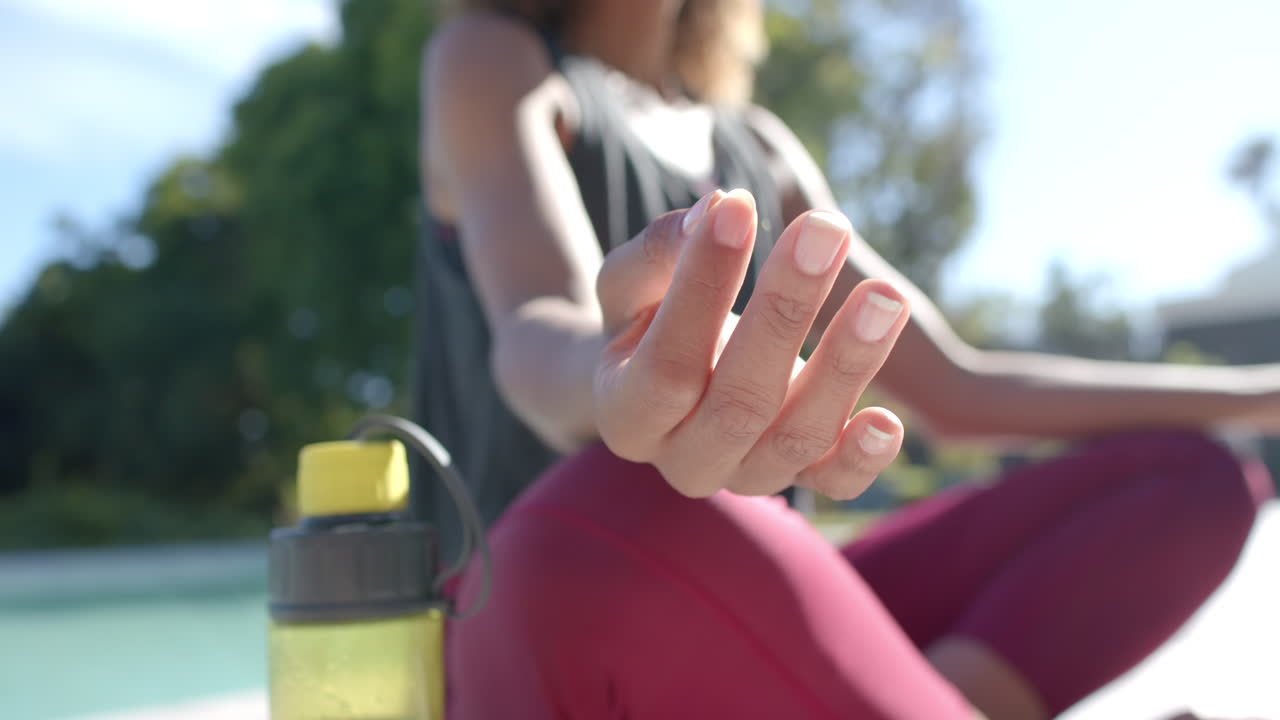 sección media de una mujer biracial practicando meditación de yoga sentada en un jardín soleado, cámara lenta