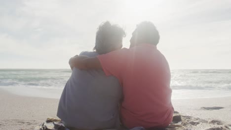 back view of happy hispanic senior couple sitting and embracing on beach at sunset