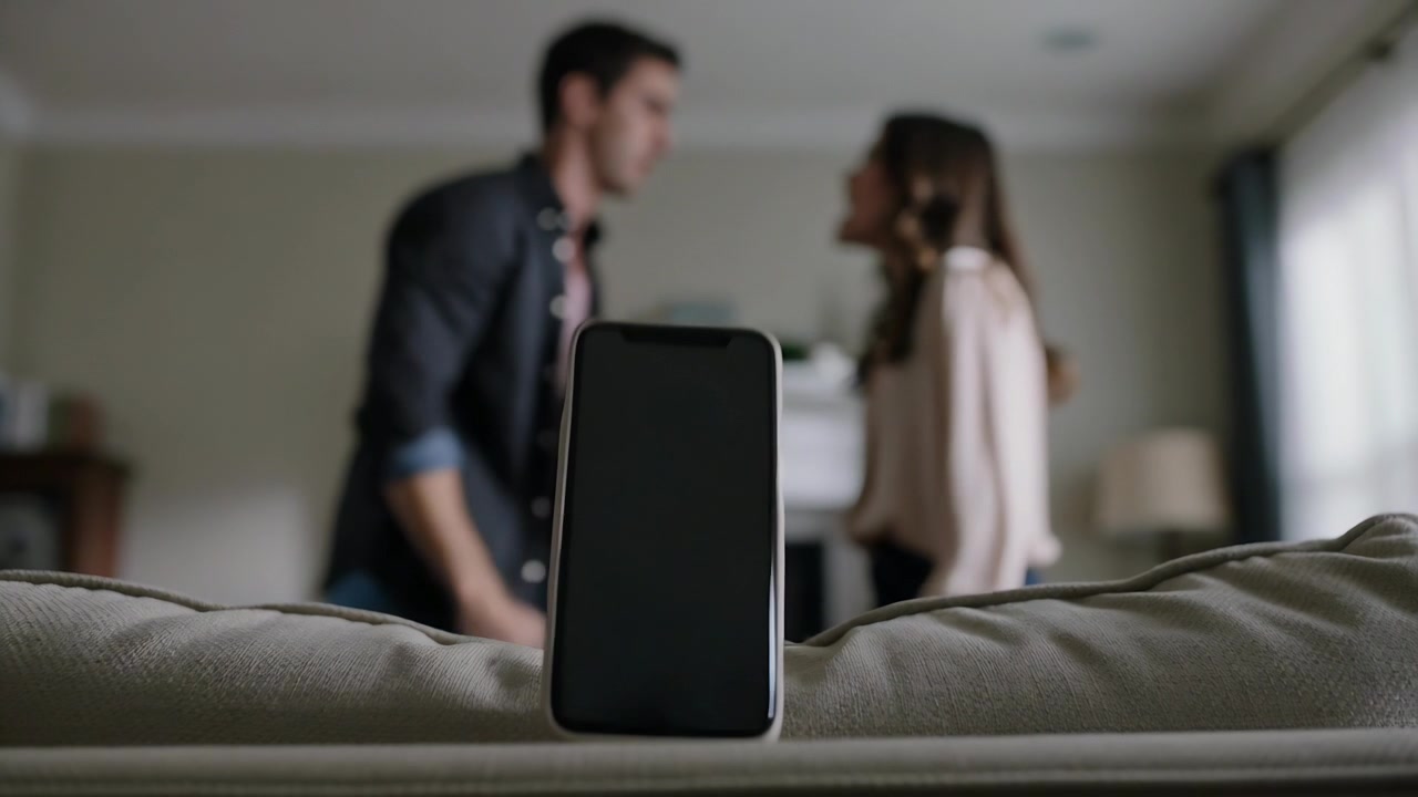 Flipping phone upright on cushion at home amid argument, man in tie, woman in blouse gesturing