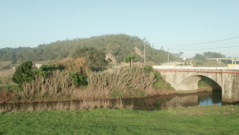 Coche-Que-Viaja-A-Través-Del-Puente-De-Arco-De-Piedra-Sobre-El-Tranquilo-Lago-Del-Río-Alcobaca-Cerca-De-Nazare-En-Portugal