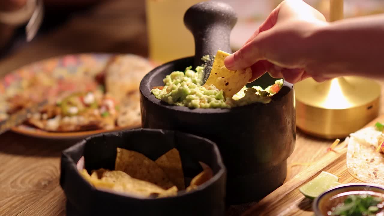 A hand dips a tortilla chip into a stone bowl of guacamole under warm, ambient lighting, with Mexican food and wooden table visible in background