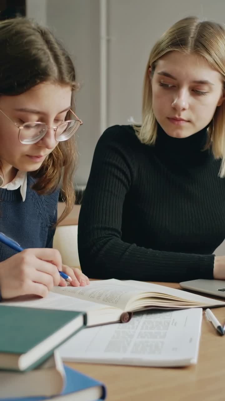 Video verticale: Studente in maglione che prende appunti su un libro aperto con una penna blu al tavolo, un compagno studia al laptop