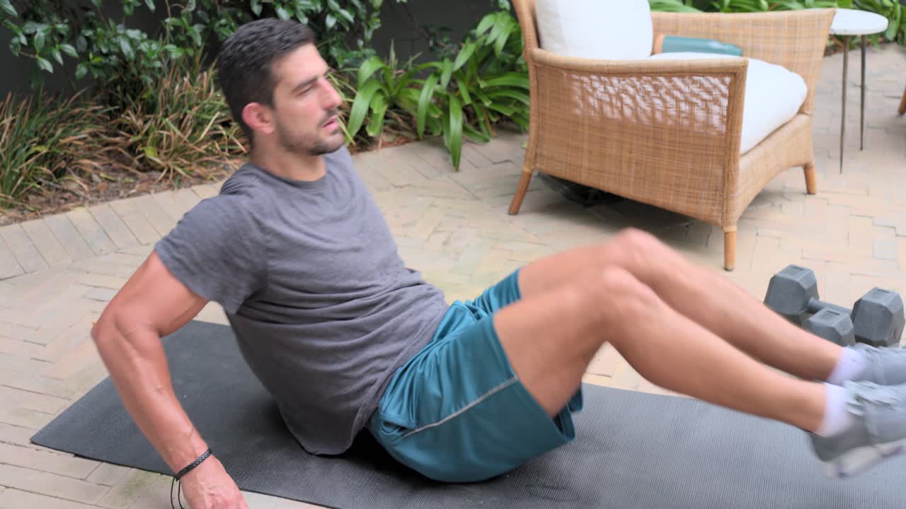 Uomo che appoggia le mani, esegue sollevamenti delle gambe su un tappetino scuro, nel patio del cortile vicino a manubri esagonali, tonificando il core.