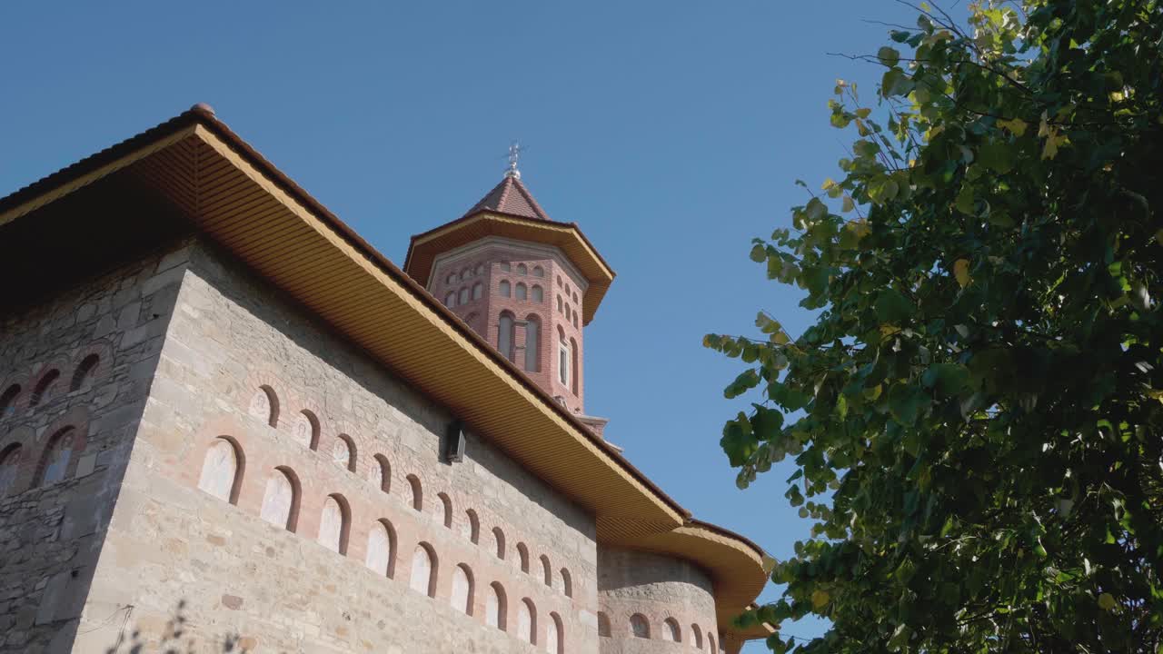 A low-angle tilting shot reveals the historic Precista Church in Bacau, Romania. The camera moves up the medieval stone and brick walls to the tower against a clear blue sky