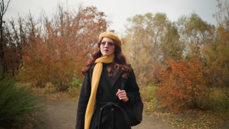 student in black coat, and beret strolls through serene autumn park, holding bag strap with focused expression, vibrant fall foliage and peaceful setting enhance warm seasonal atmosphere