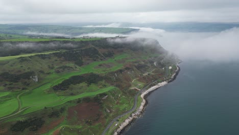 Ascending-aerial-shot-over-rocky-coast-road-near-glenarm-town-in-northern-ireland-with-view-of-sea-coast-with-blue-sea,-gorgeous-green-landscape-and-rocks