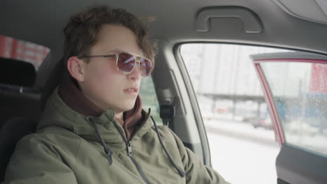 young boy enters parked car wearing winter jacket while snowy city street and passing vehicles are visible through open door, capturing urban lifestyle and seasonal atmosphere from interior view