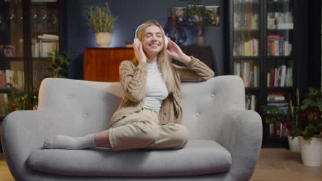 happy young woman, with closed eyes, listening music in wireless headphones while sitting on a couch at home