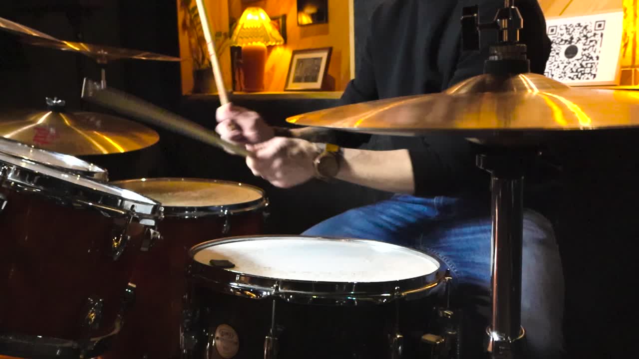 Slow motion footage a Caucasian white man with tattoos and blue jeans playing acoustic drums indoors in a rehearsal room by hitting cymbals, hi-hat and snare drum hard. Lamps with warm light in back.
