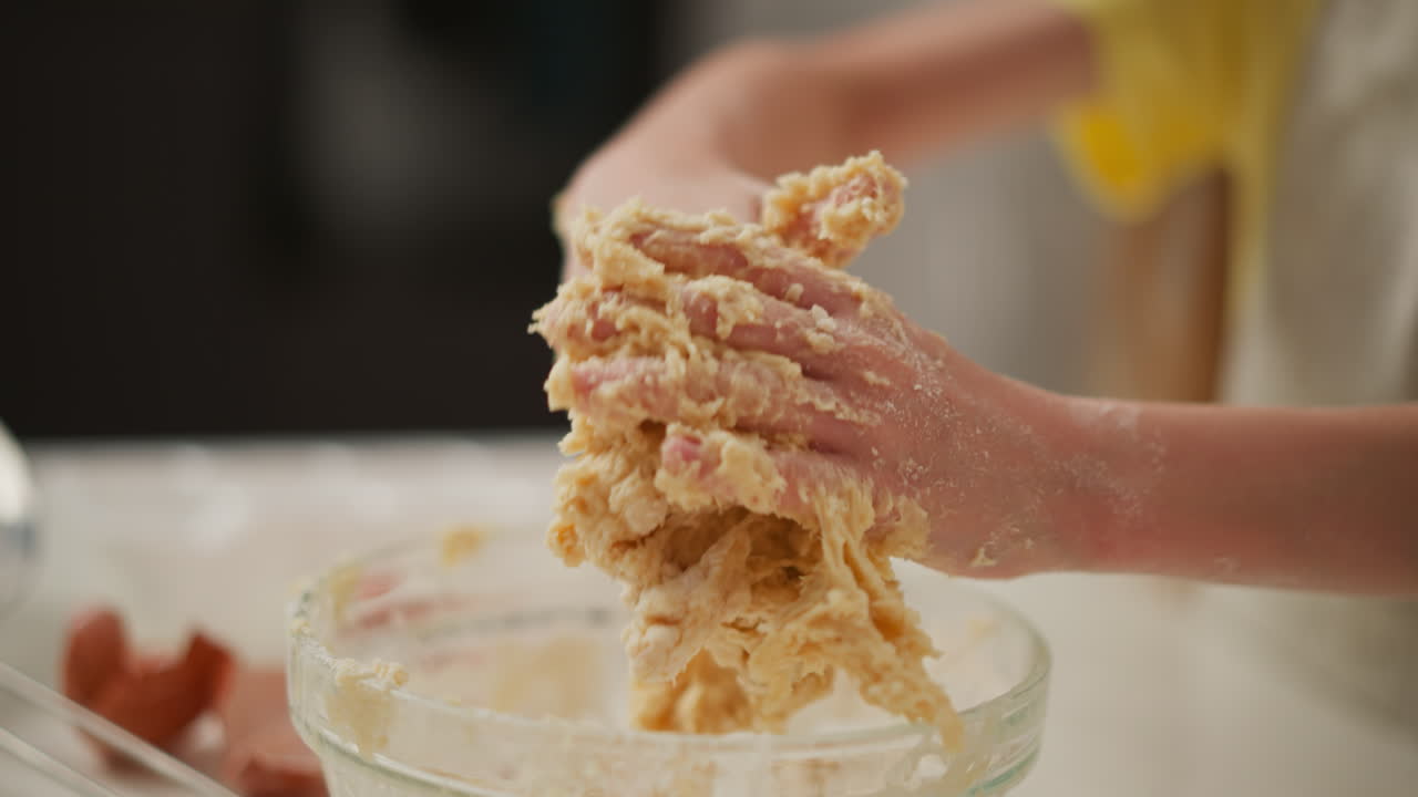 Close up of person kneading sticky dough in glass bowl with blurred view of another person walking in and sprinkling flour on hand as first person continues baking messy mixture in cozy kitchen
