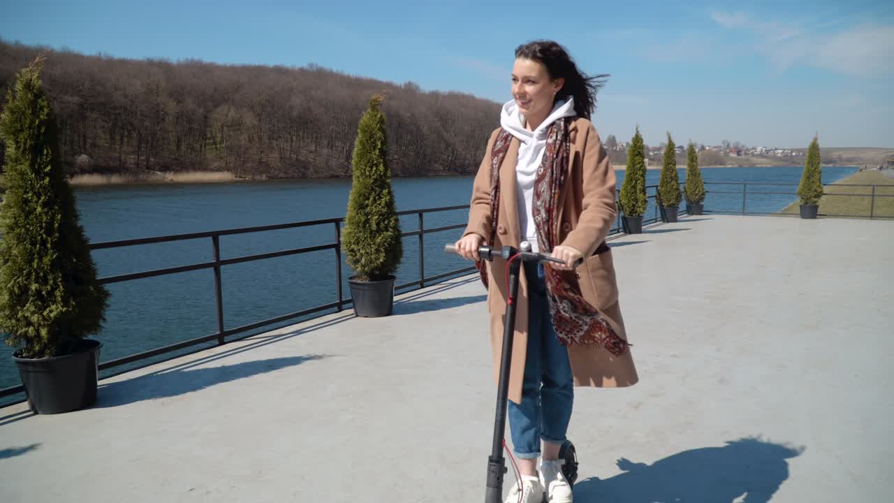 Young girl student in a sweater, jeans and a coat rides on an electric scooter in a beautiful area near the river. Front view. Ecological city transport