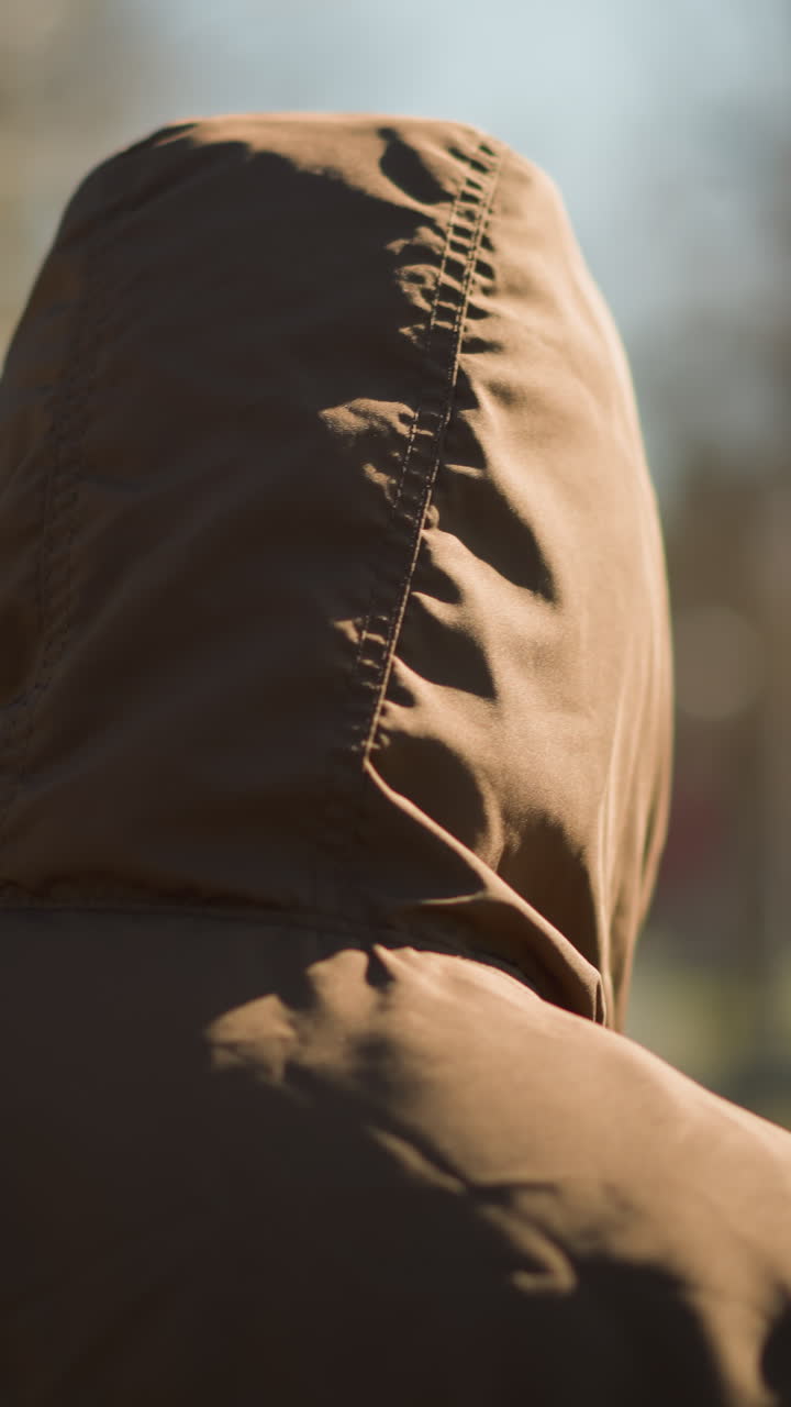 un primer plano de un individuo con una chaqueta con capucha marrón caminando al aire libre, con un fondo borroso de árboles y un camino