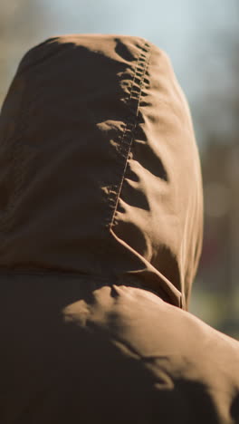 a close- up of an individual wearing a brown hooded jacket walking outdoors, with a blurred background of trees and a pathway
