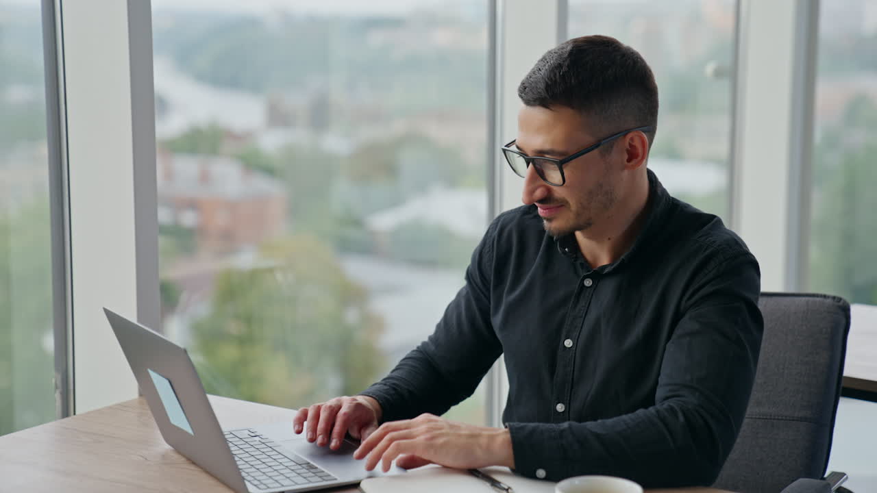 Dark-haired man freelancer working at laptop. Relaxed man in a good mood working at the desk on his computer. Blurred backdrop.