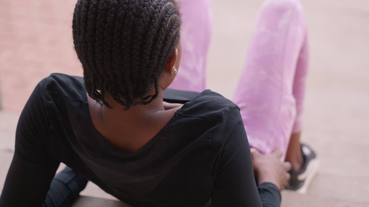 Adolescente di colore reclinata su scalini esterni con pantaloni rosa e capelli intrecciati, top nero casual, postura rilassata appoggiata all'indietro, luce diurna soffusa, scalinata urbana, ritratto spontaneo di gioventù che trasmette calma e cura di sé.